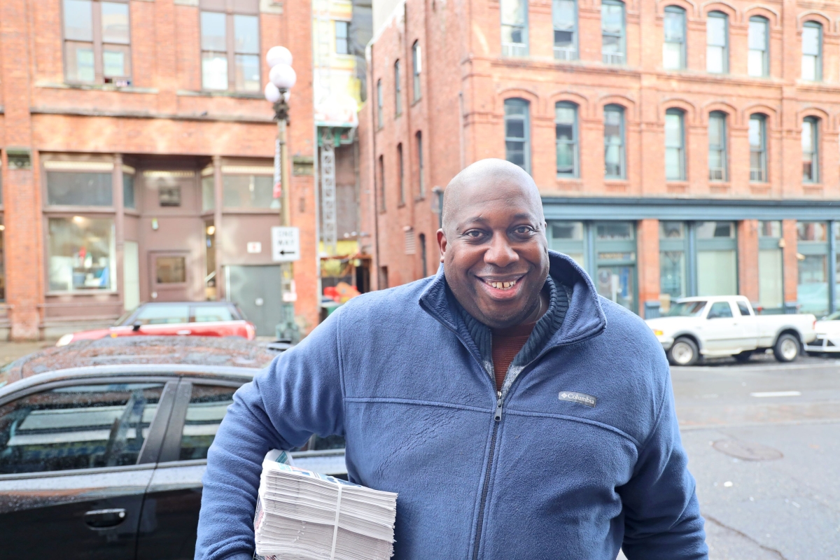 Smiling Black man in fleece jacket stands on city street holding a stack of newspapers in one arm.