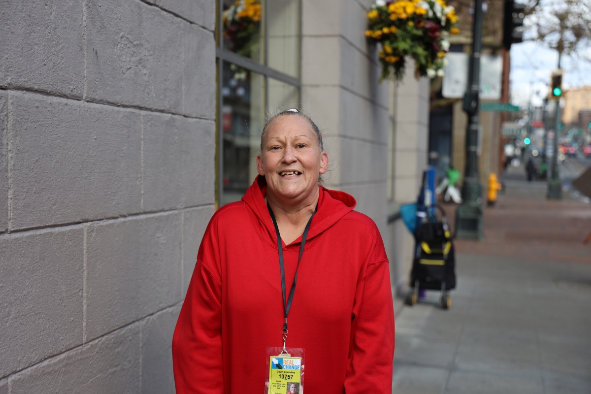 A smiling woman stands on the sidewalk. She has a Real Change badge on.