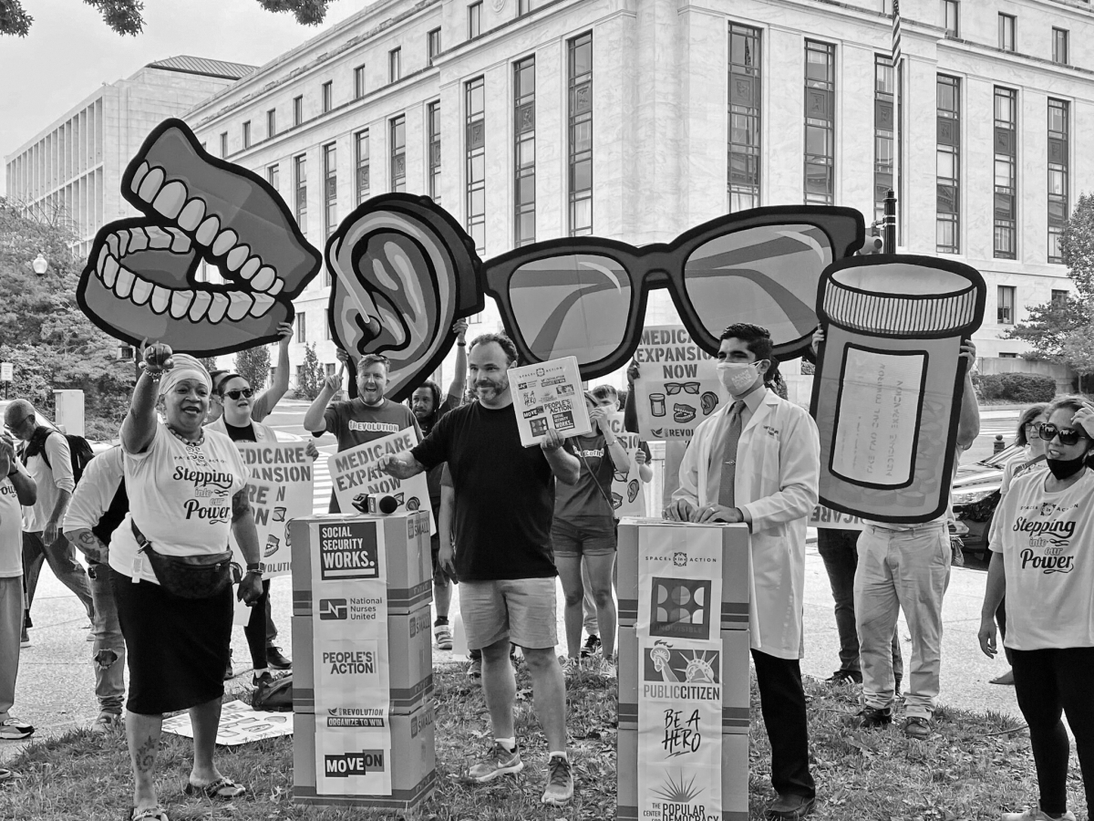 Protesters outside government building holding signs and large models of teeth, ears, eyeglasses, and prescription bottle
