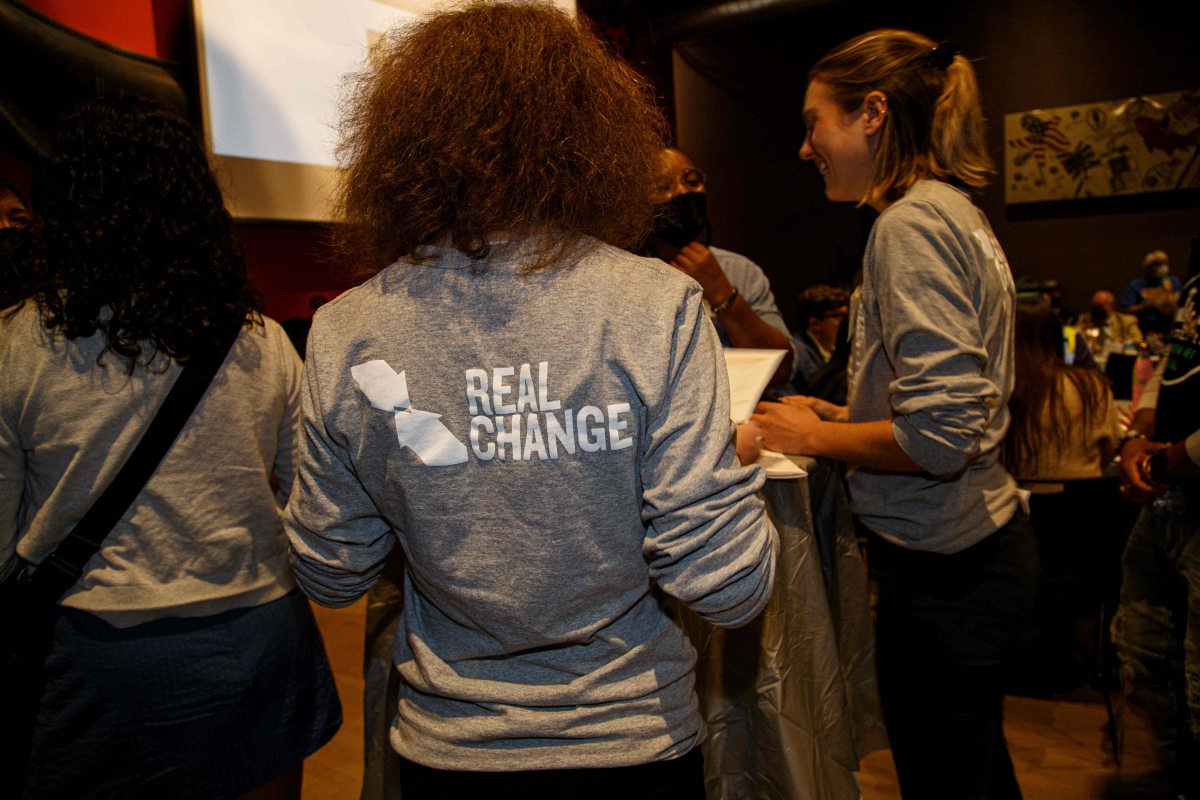 Young women in Real Change shirts, seen from behind, stand in office space.