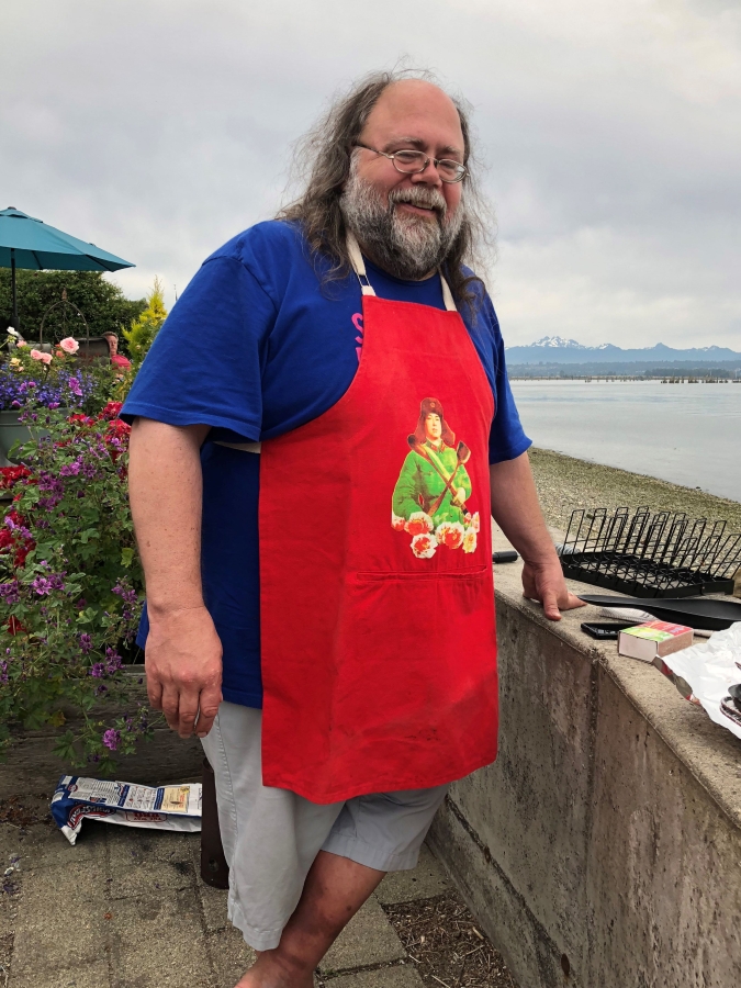 Scott Morrow standing in an apron at an outdoor grilling party with an open body of water in the background