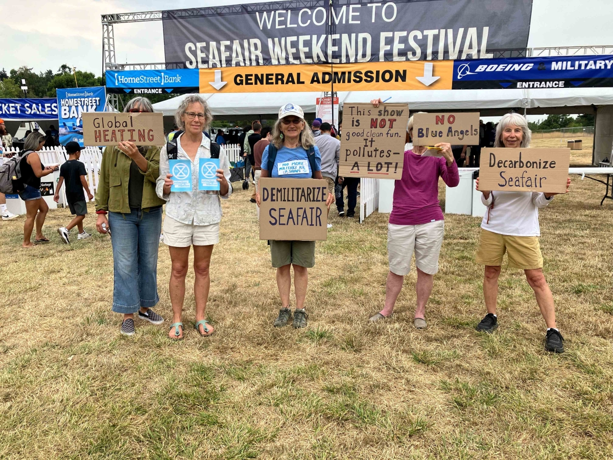 Six people stand on Seafair Weekend Festival grounds holding signs with messages such as, "Demilitarize Seafair" and "Decarbonize Seafair."