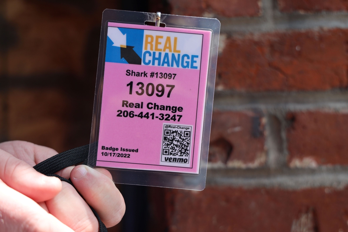 Photograph of hand holding pink badge, showing number 13097, against backdrop of brick wall