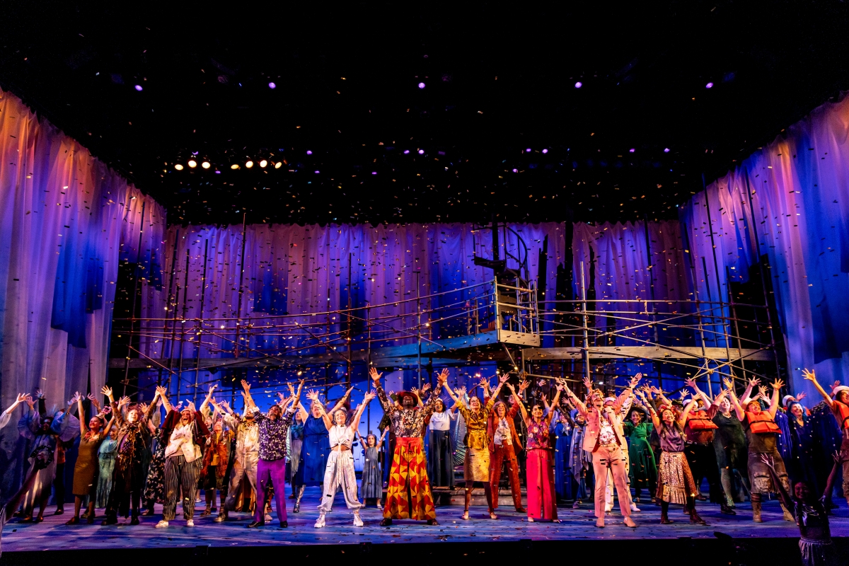 Large cast of colorfully dressed people stand with legs apart and arms raised on a lit-up stage against a purple background