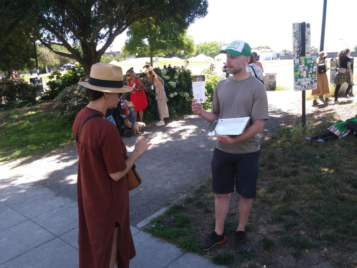 A woman in a sunhat and red dress talks with a man wearing cap, shirt, and shorts and holding a clipboard.