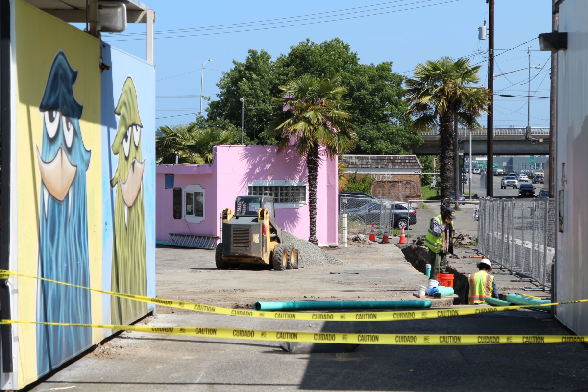 Photograph of lot on sunny day, yellow caution tape visible in foreground and pink building in background, with view of murals on one side and workers in vests on the other