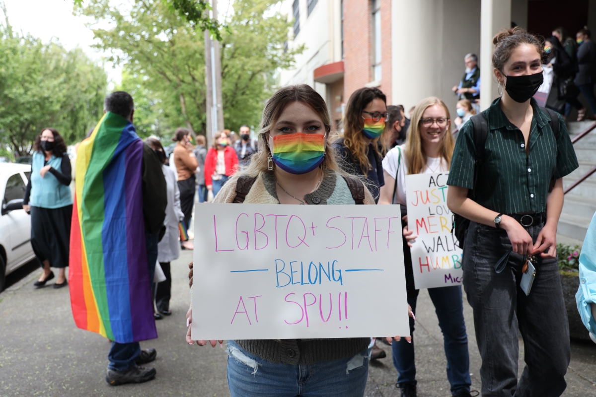 Several masked young people walk in rally; young woman in rainbow mask in foreground carries sign reading, "LGBTQ+ Staff Belong at SPU!!"