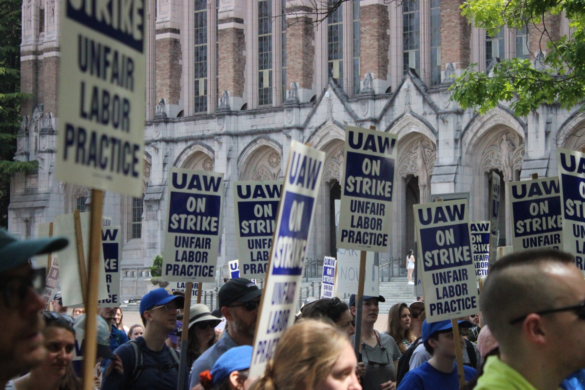Crowd of people outside on sunny day carrying signs saying, "UAW On Strike."