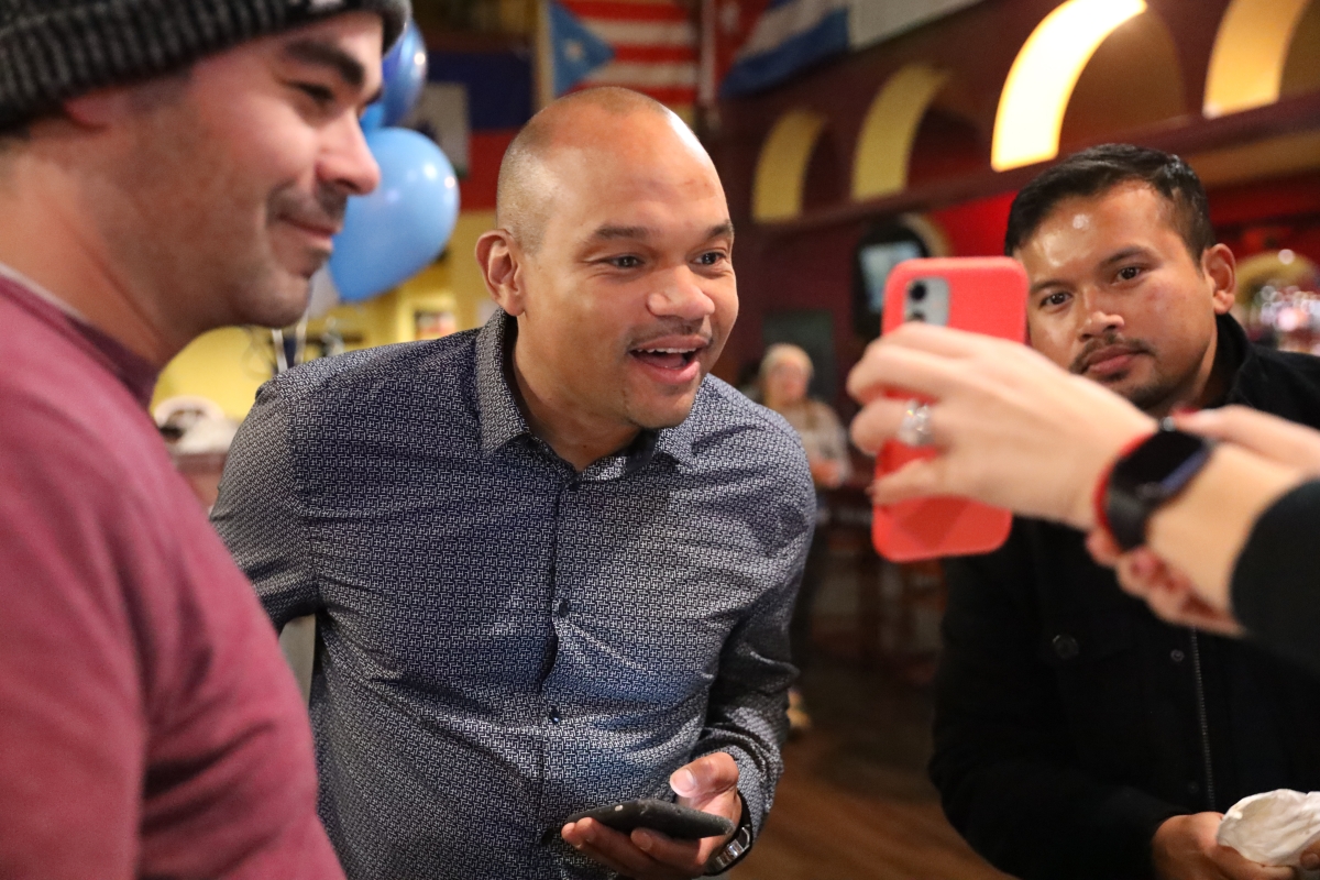 Chipalo Street, dressed in blue-and-white print shirt, flanked by two young Latine-appearing men, smiles into a phone screen being held up to his gaze