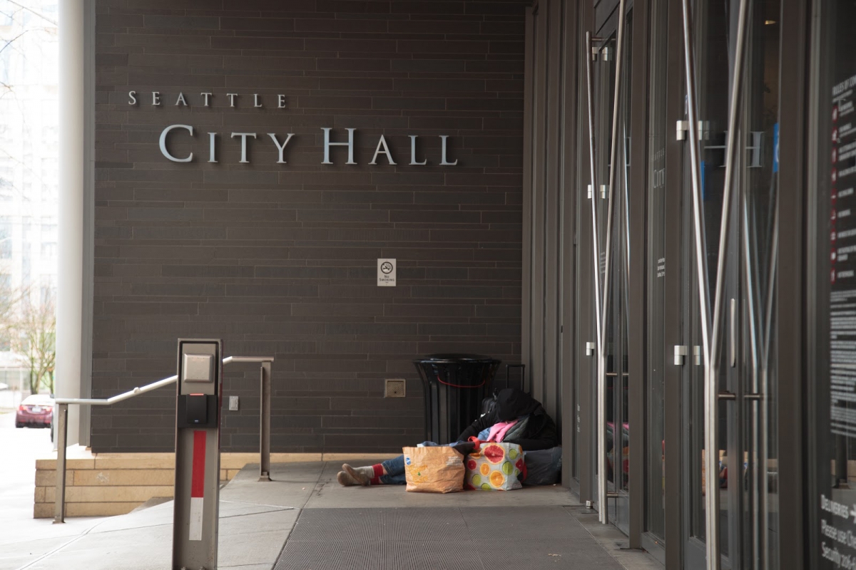 Entrance to Seattle City Hall. A person seen in profile, sitting next to a trash can, surrounded by belongings in plastic bags