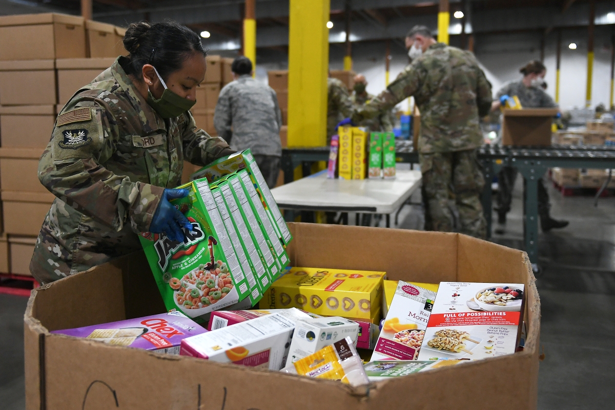 Photograph of young Black woman in National Guard uniform and face mask lifting boxes of cereal from a box in storeroom with other uniformed Guardspeople visible in background