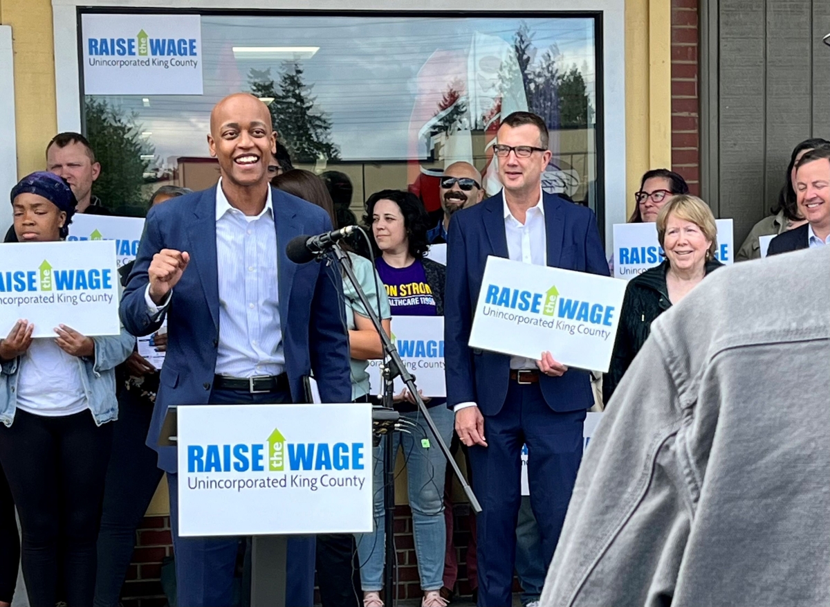 Girmay Zahilay, young Black man in suit jacket, stands at a microphone and podium with a sign reading, "Raise the Wage," backed by several other people in front of a glass window.  