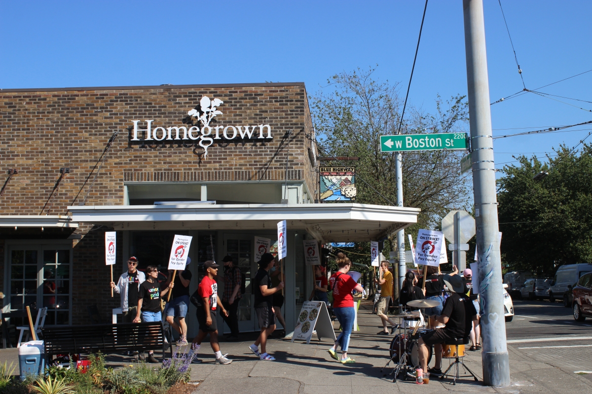 Picketers walk outside Homegrown store on sunny day. Street sign reads, W. Boston St.