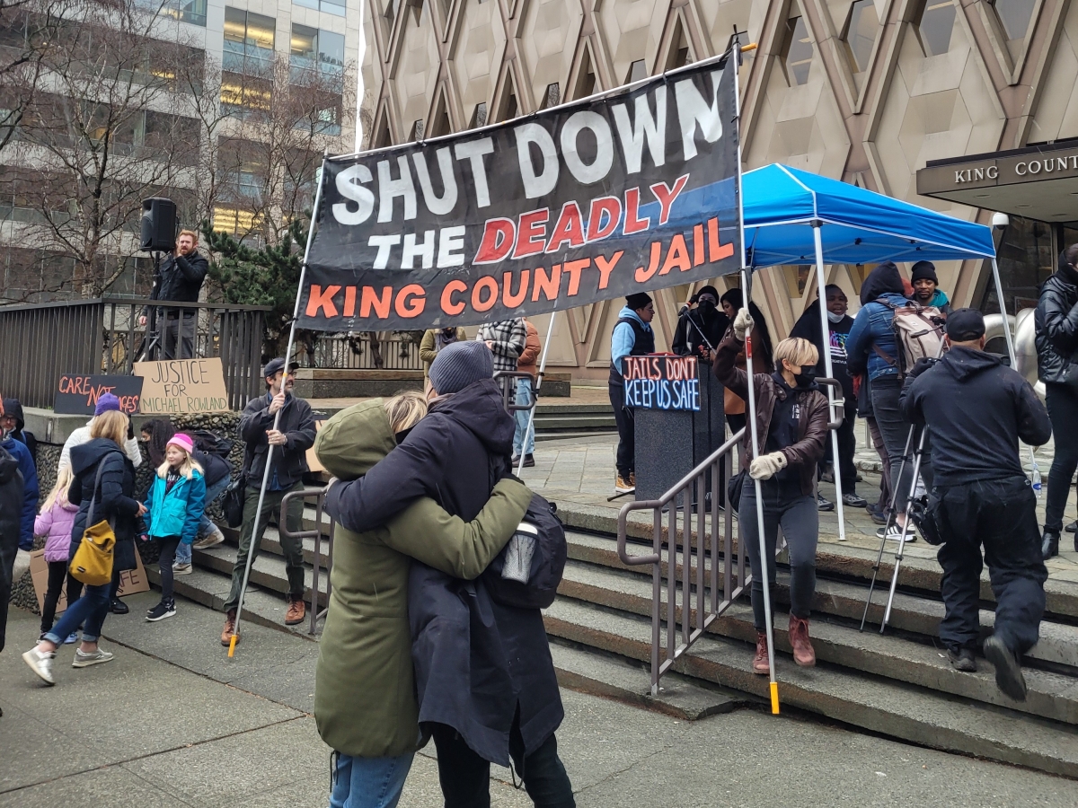 People in coats and hats, two of them embracing, stand outside King County jail, under banner that reads, "Shut Down the Deadly King County Jail."