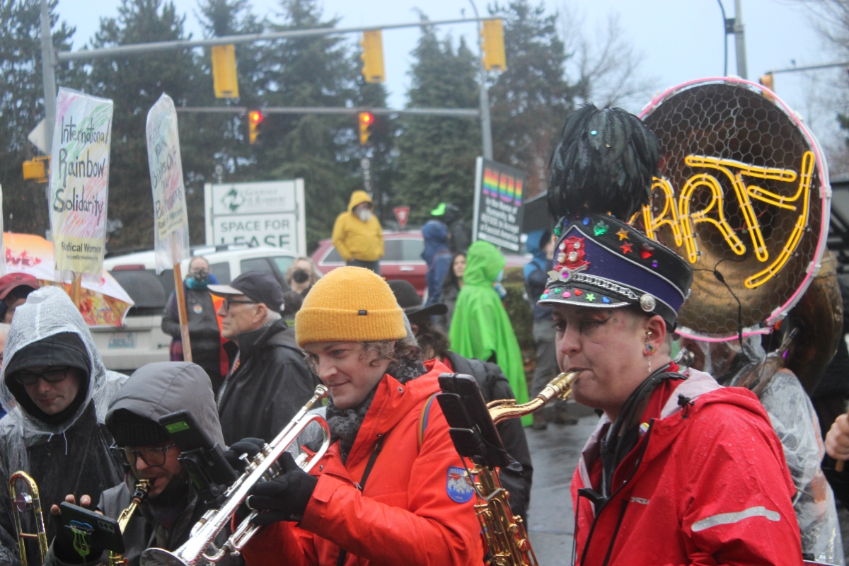 Four people in hats and hoods, one in a bandmaster's cap, play wind instruments at head of crowd in an outdoor setting.