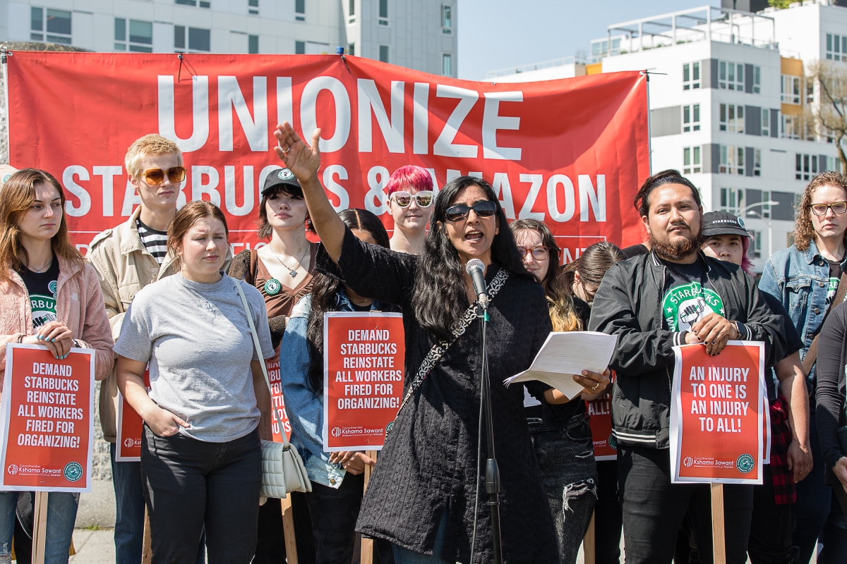Kshama Sawant stands in front of sign reading, "Unionize Starbucks & Amazon" among several young people holding pro-worker signs.