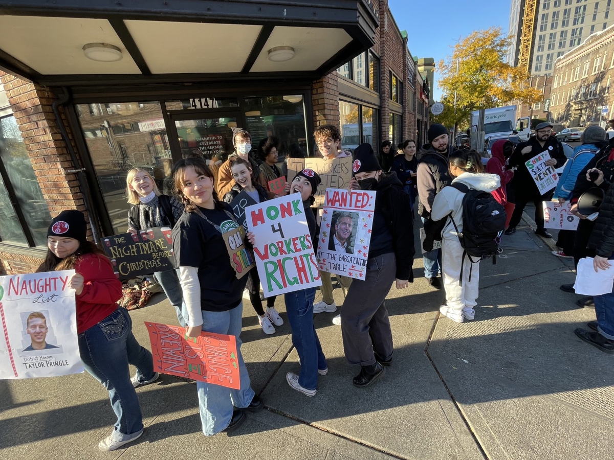 Large group of smiling young people holding picket signs gather on street corner outside a Starbucks shop on a sunny day.