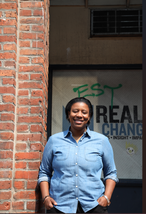 Joy Hollingsworth, young Black woman in button-down flannel shirt, stands by brick wall with Real Change banner visible in background.