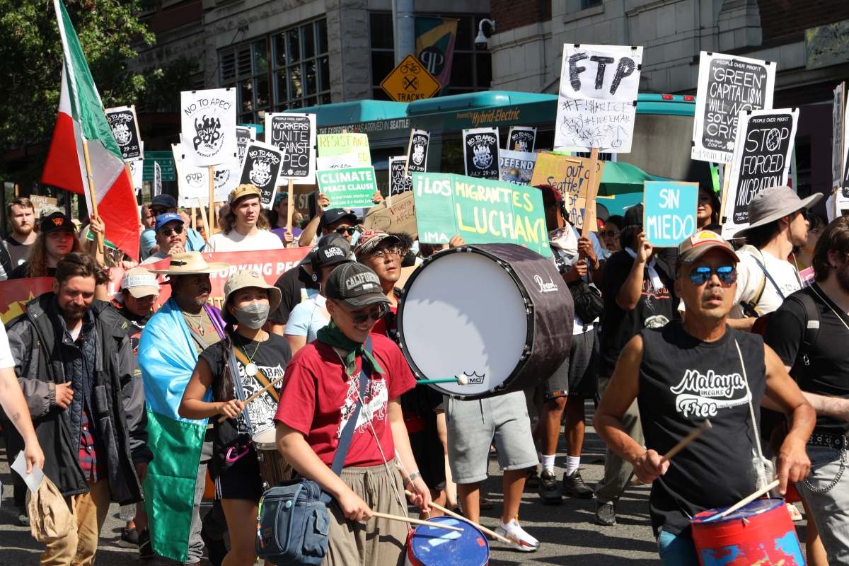 A large group of people fill a city street with drums and signs on a sunny day.