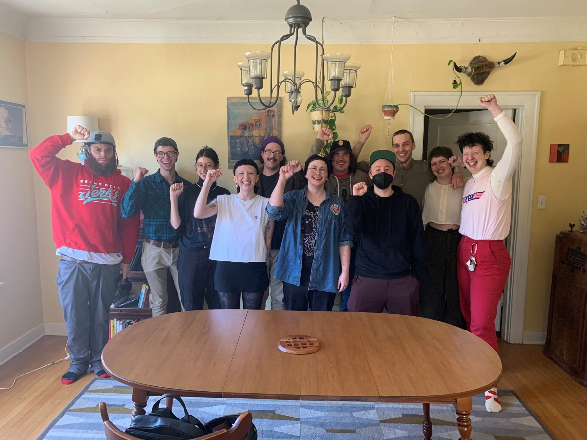 Photograph of group of young people, of various races and genders, many wearing caps, standing in a living room behind a table with fists raised in salute