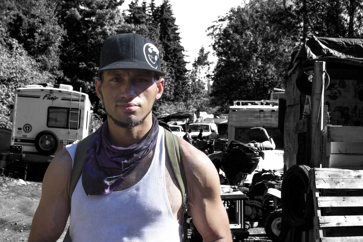 A photo of north Seattle encampment resident Robert Barden in a white tank top with a hat and bandana on. Barden is colorized and the encampment behind him is in black and white, symbolizing that he is a person first.