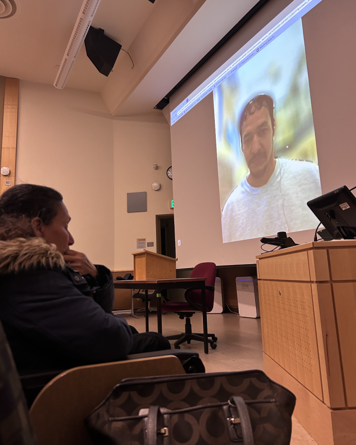 Person in jacket with parka collar sits in chair in a classroom; a man appears on screen projected on wall.