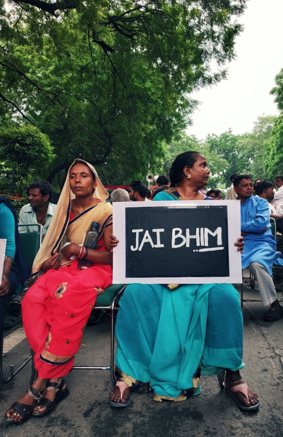 Two women in saris sit next to each other in outdoor meeting; one holds a sign that reads, "Jai Bhim."