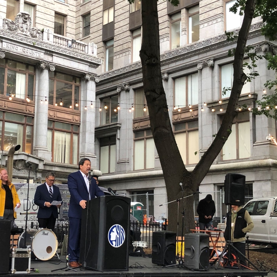 Bruce Harrell stands at a podium under a tree in front of a building.