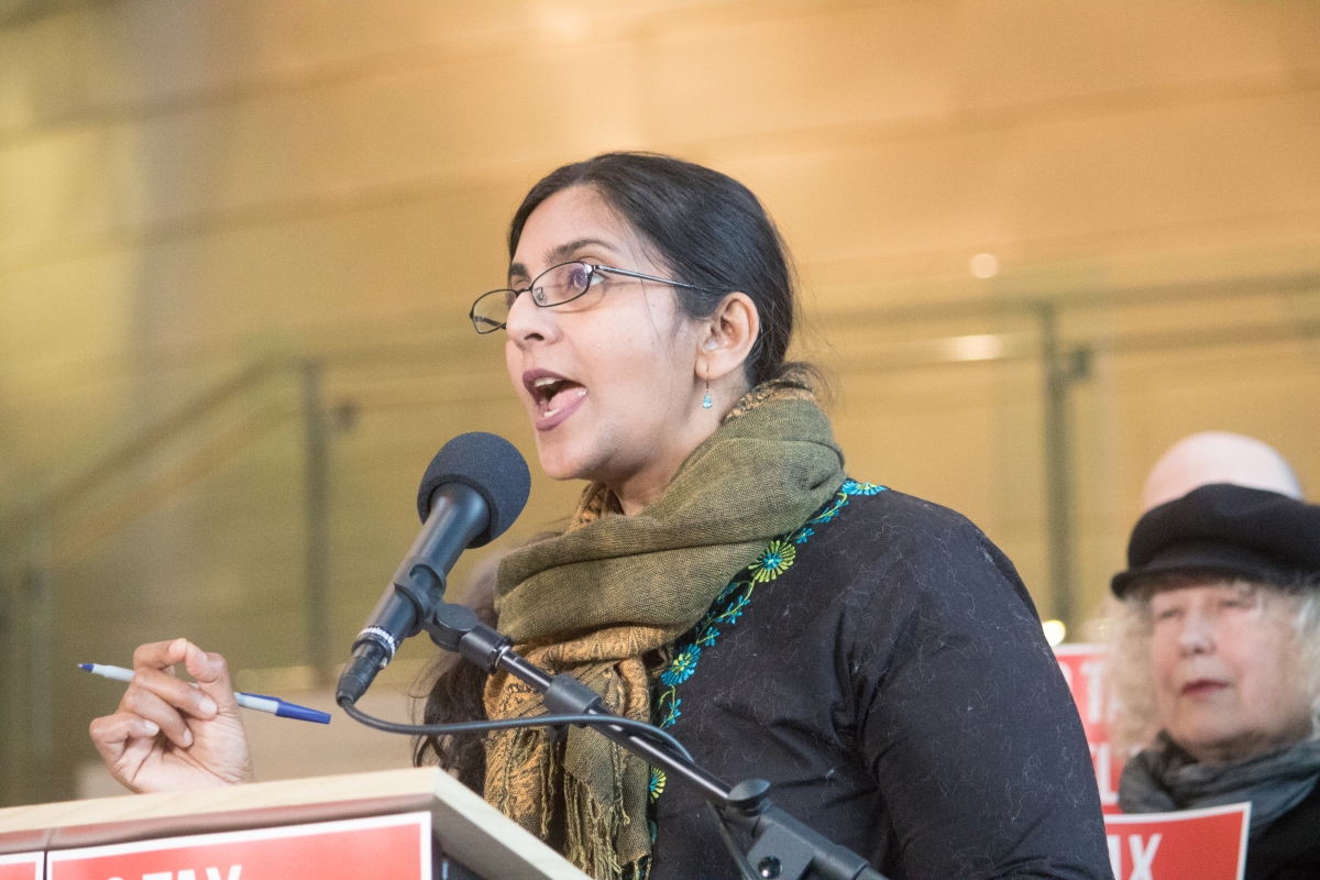 Woman wearing glasses, earrings, and neck scarf stands at a podium and speaks, holding pen in right hand.