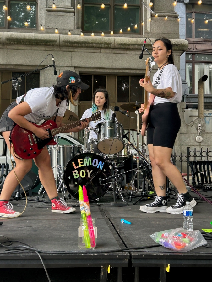 A trio of young women on stage in a public park play various insturments, including a base, a drum set and a guitar.