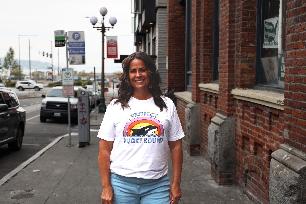 Maren Costa, young Latina woman with shoulder-length brown hair, dressed in white T-shirt reading, "Protect Puget Sound," stands on city street by red-brick Real Change building.