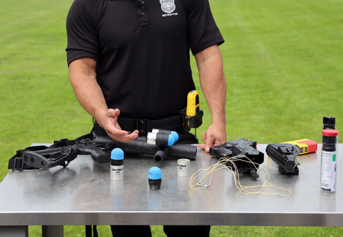Photograph of torso and arms of uniformed officer handling various weapons, cords, and capsule bottles arrayed on a table