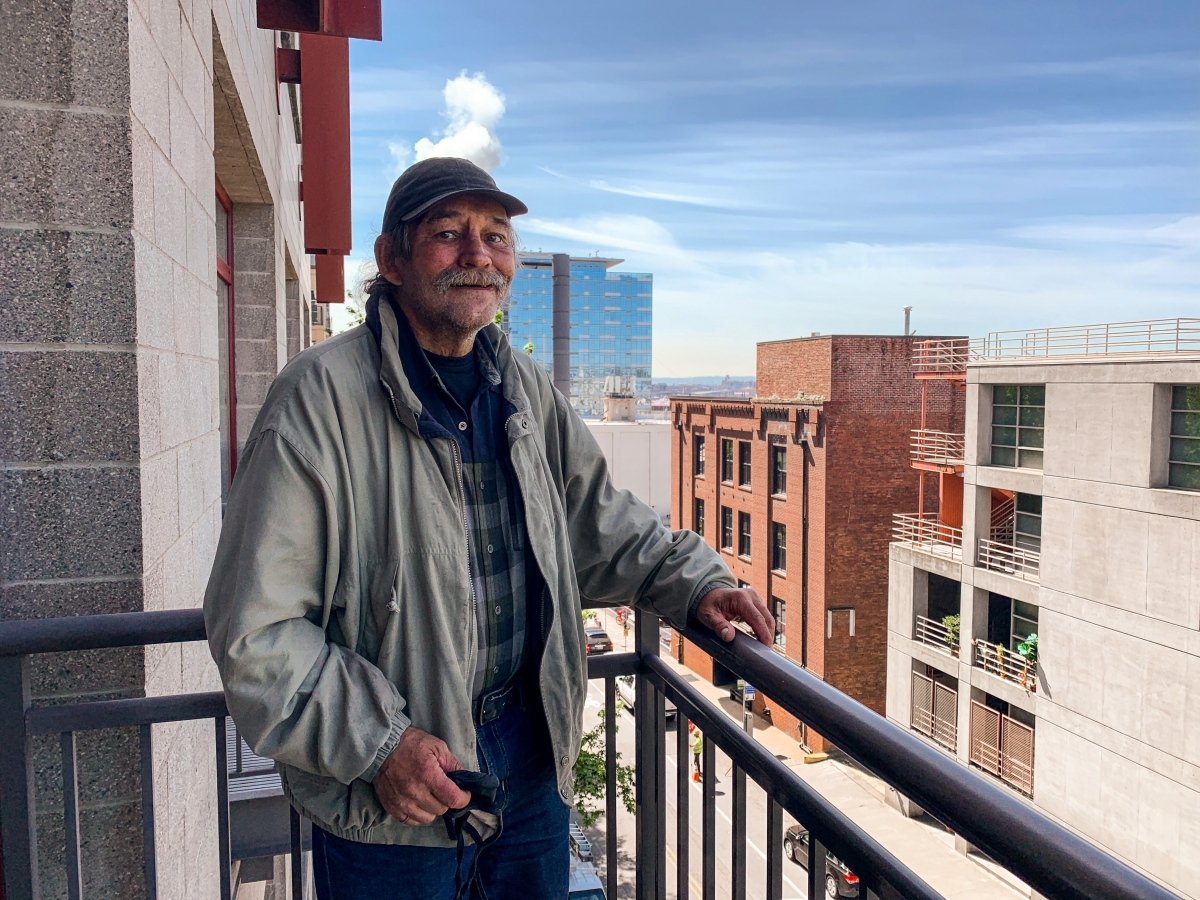 Man in late middle age with short-cropped mustache and beard, in cap and light-colored jacket over flannel shirt, standing on high-rise balcony with buildings in background