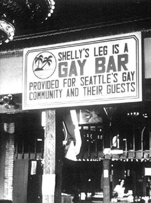Photograph of front window of bar with sign reading, "Shelly's Leg Is a GAY BAR..." next to a logo featuring two palm trees