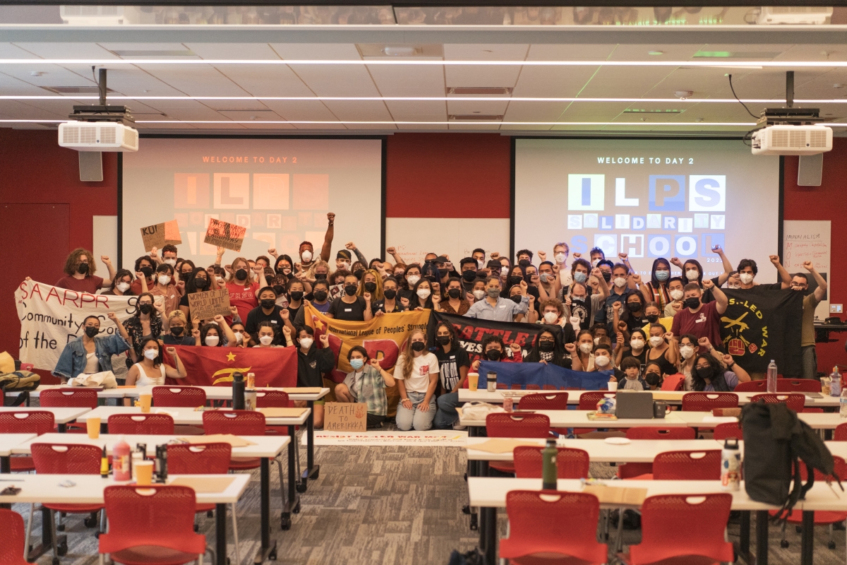 Large group of young people at the front of a classroom, backed by screens reading, "Welcome to Day 2--ILPS," pose for camera holding banners and raising fists.