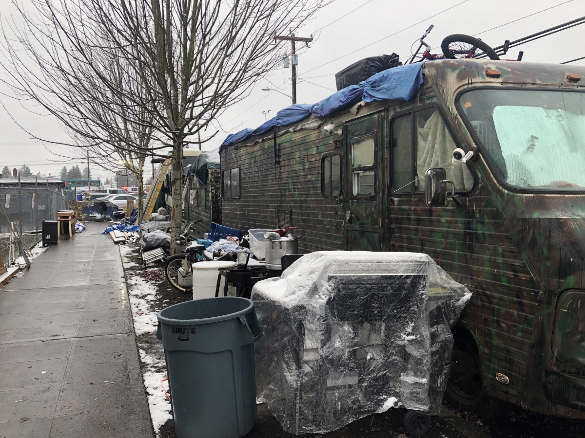Two large, green-stained RVs parked along slushy city curb on gray day, flanked by bicycle, bin, plastic-covered grill, and assorted other furniture and trash