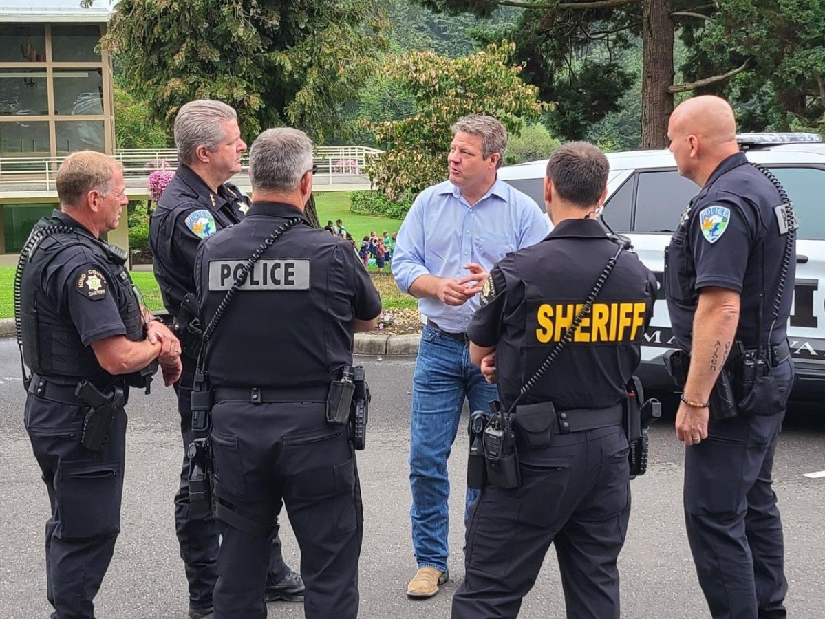 County Council member Reagan Dunn, in blue shirt and jeans, facing camera, stands speaking with five men in sheriff and police uniforms, their backs to camera.