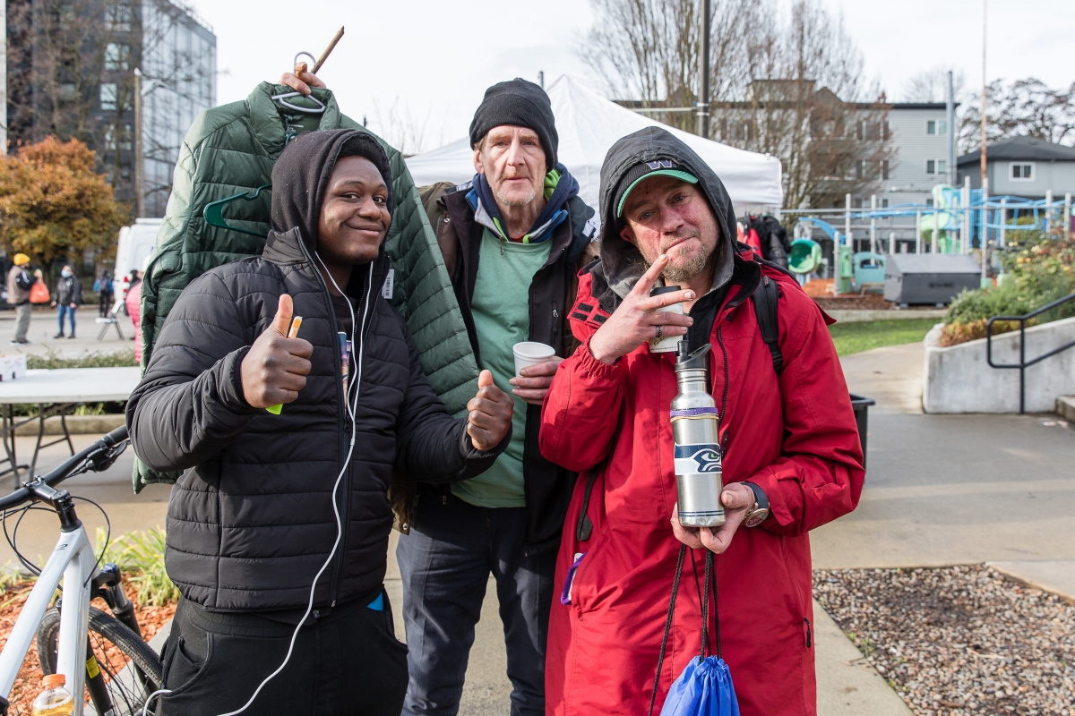 On a city sidewalk, a young Black man gives thumbs up; older white man next to him, holds up a green jacket on a hanger; middle-aged white man, wearing red jacket, on far right, holds thermos in one hand and makes rap/peace sign with the other. 