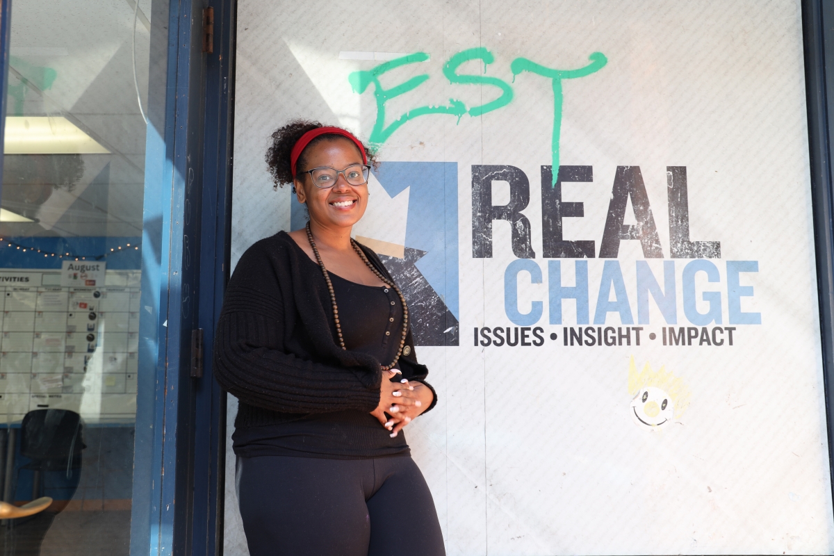 Young Black woman wearing glasses, dark sweater, and beaded necklace stands in front of Real Change banner