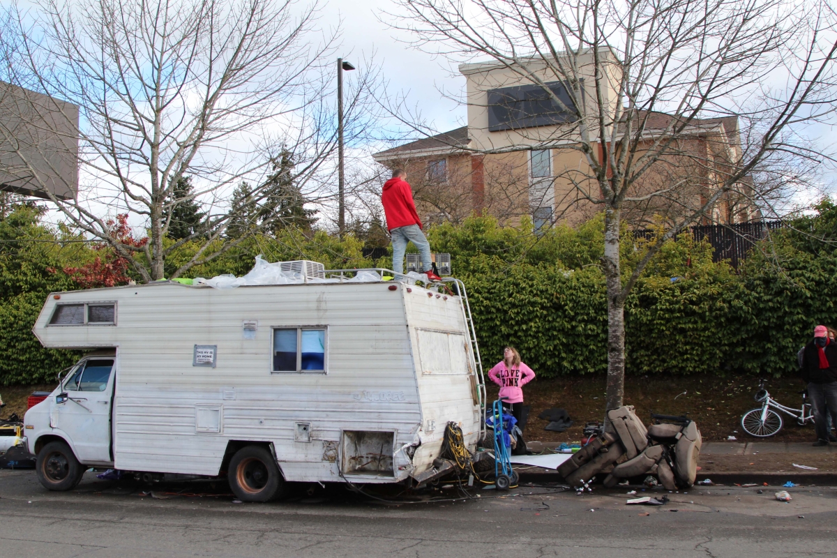 Man stands on top of RV while woman stands on sidewalk looking up at him. 