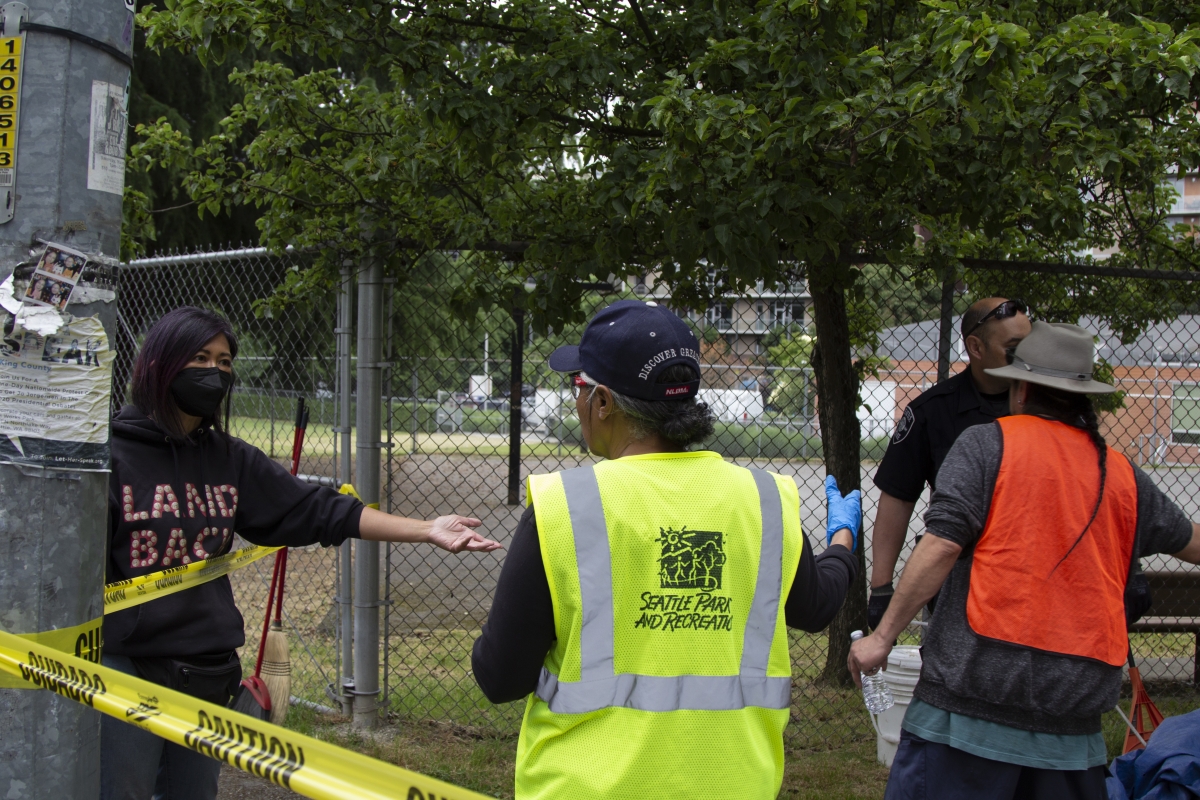 A stop the sweeps volunteer wearing a "Land Back" hoodie yells in exasperation at a city cleanup worker in a bright yellow vest during an encampment removal, while the camper, with his braided ponytail sticking out from under a brimmed hat, looks on.