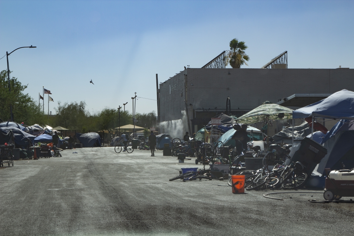Photograph of large lot with windowless, block-size building in background, tents and bicycles on perimeter in foreground and background