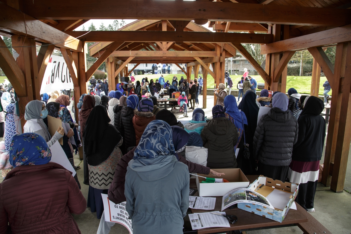 Crowd of women in jackets and hijabs, seen from behind, standing outside under wooden girders.