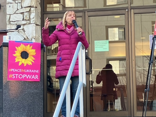 Medea Benjamin, middle-aged white woman with shoulder-length light hair and wearing purple quilted jacket, stands at top of steps holding a microphone, next to a sign calling for peace in Ukraine.