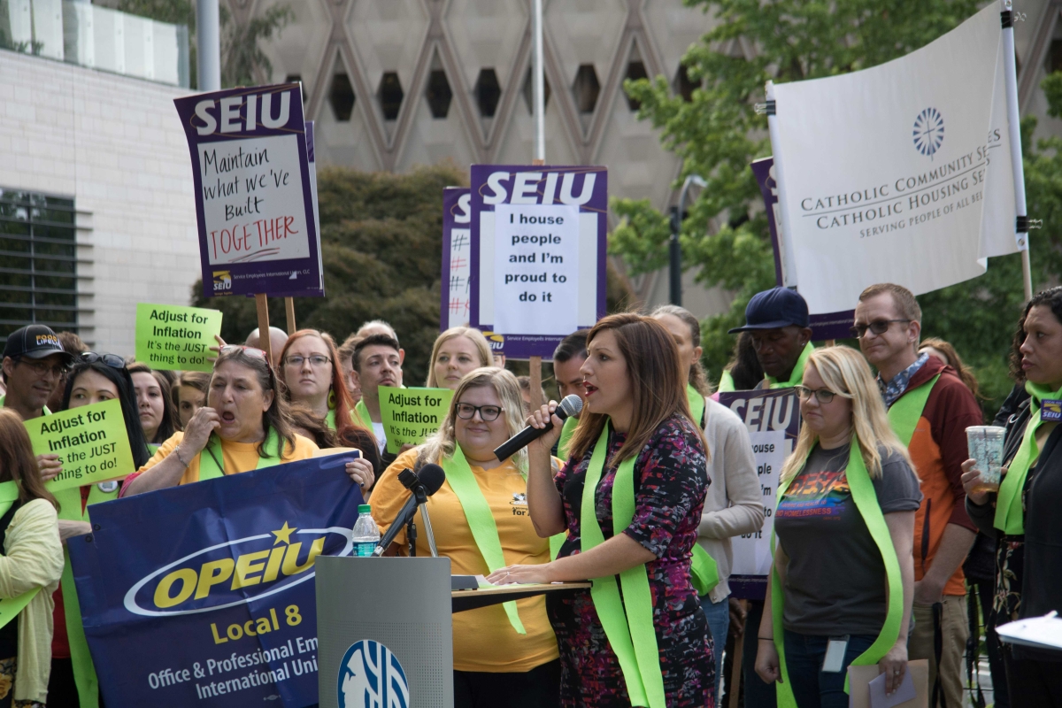 Teresa Mosqueda stands at podium, speaking into a microphone, in outdoor setting, surrounded by people holding green SEIU signs.