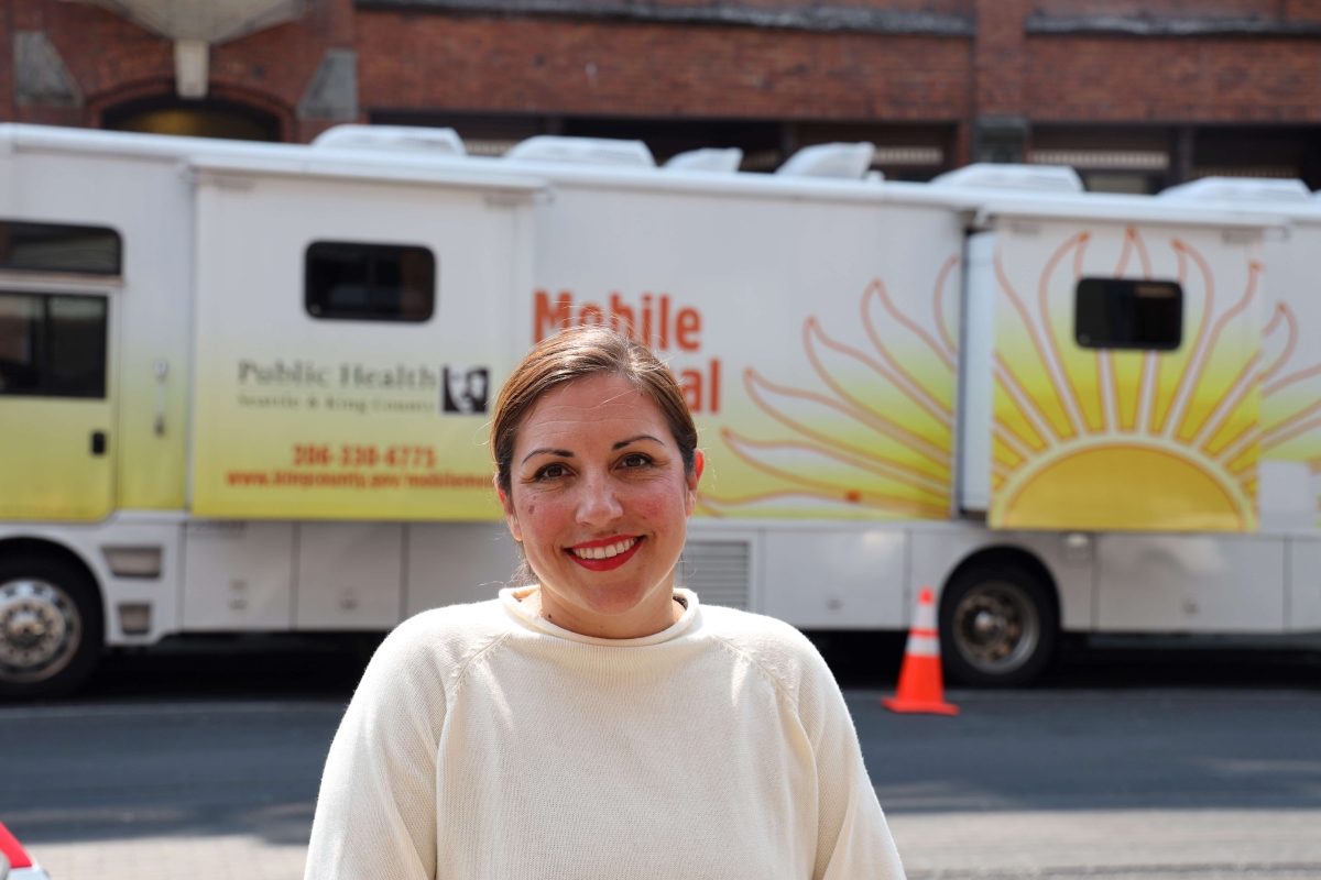 Teresa Mosqueda, young to middle-aged Latina woman dressed in white sweater and with hair pulled back, stands on street on sunny day with mobile Public Health vans visible behind her.