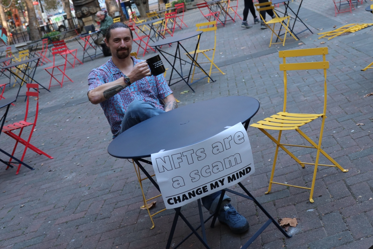 Photograph of Tobias Coughlin-Bogue, young white man with dark hair in ponytail and mustache and beard, sits at a metal outdoor table holding up a coffee cup; sign on table reads, "NFTs are a scam. Change my mind."