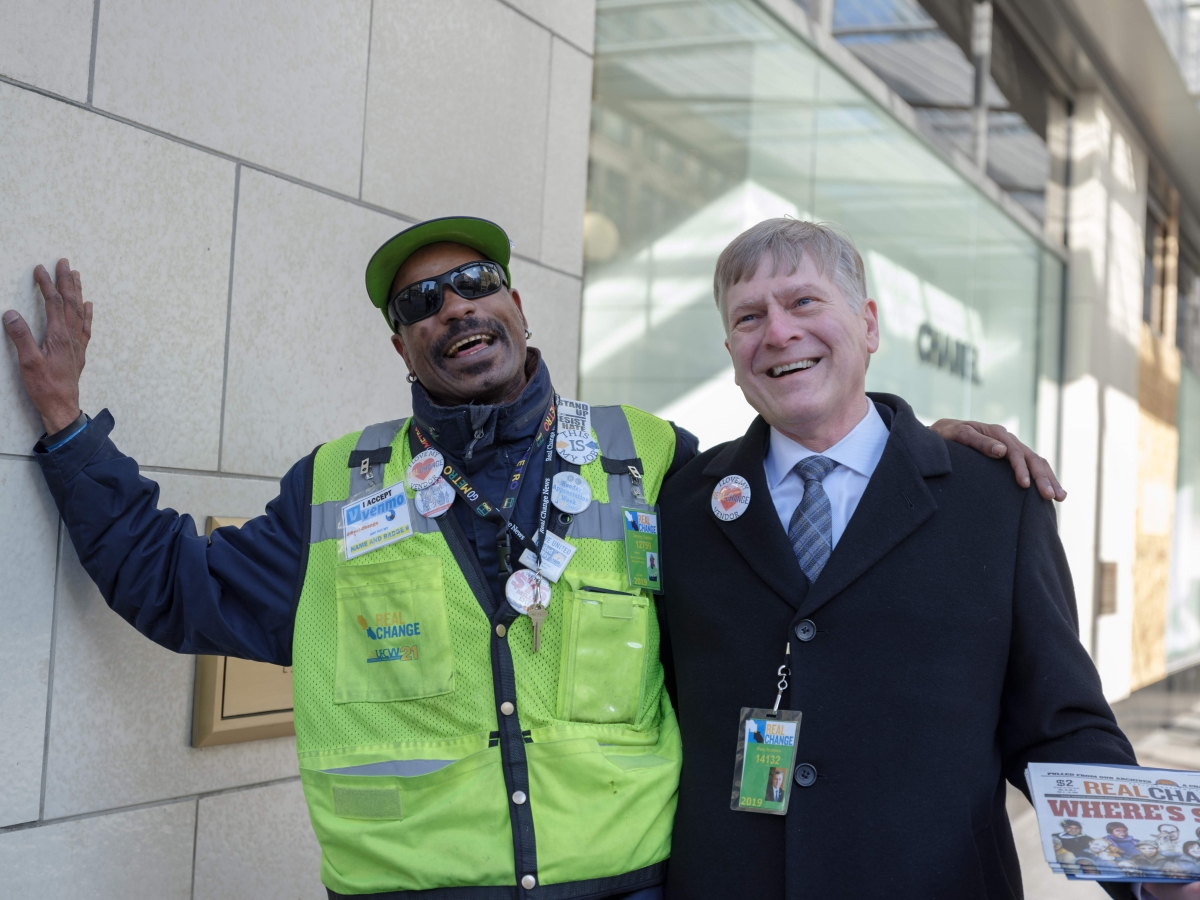 Black man in green vest stands with arm around white man in suit with green badge