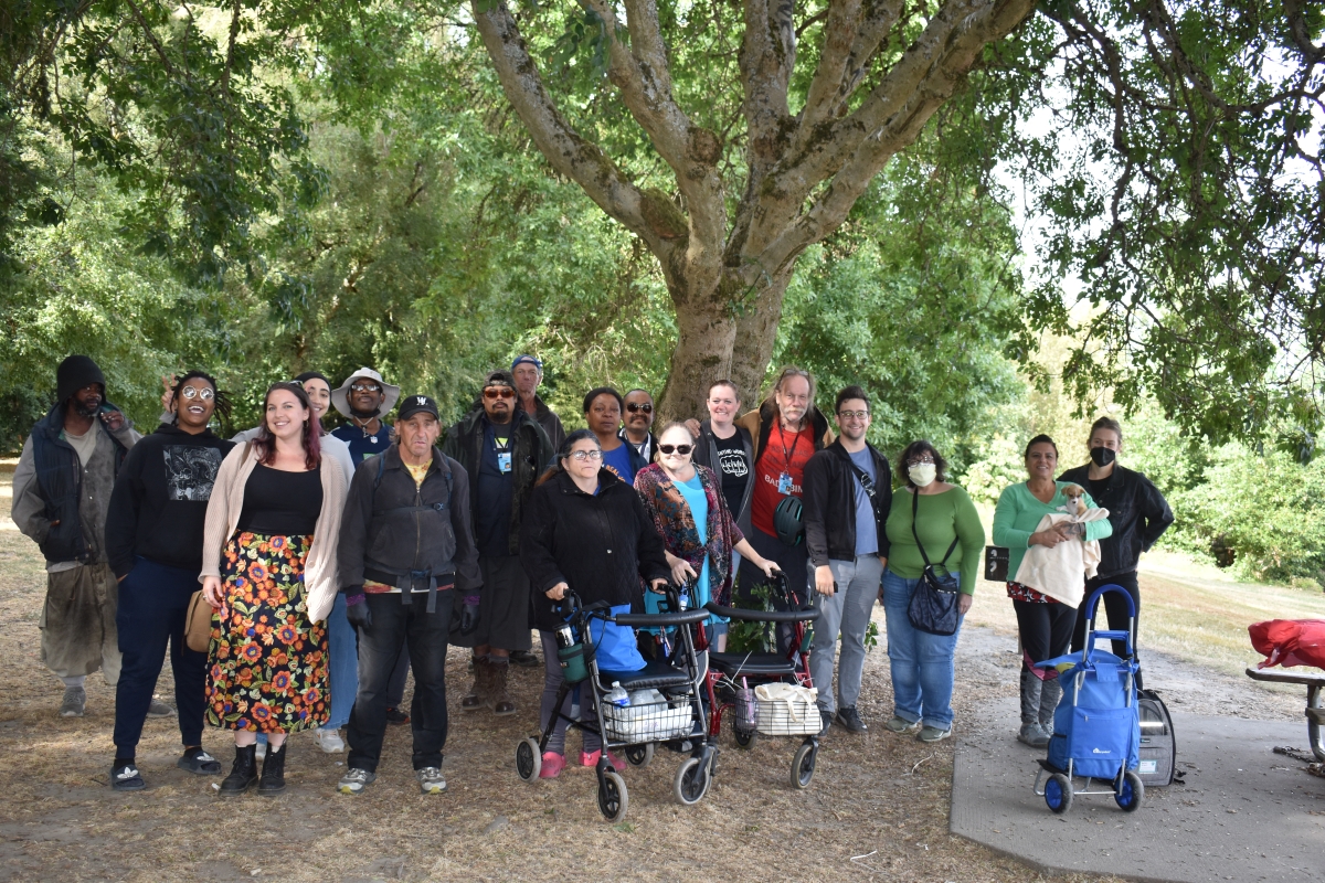 Large group of people, some in masks, some pushing carts, pose for picture in wooded outdoor setting