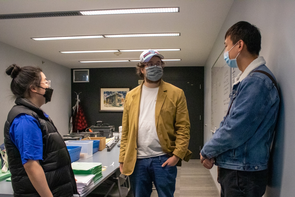 Three people in masks stand talking in a fluorescent-lit office.